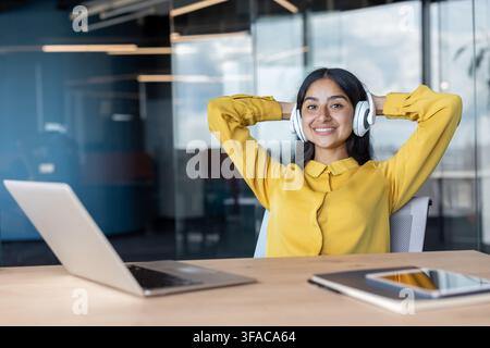 Portrait d'une jeune femme indienne portant des écouteurs assise à un bureau dans un bureau, les mains derrière la tête, se reposant et souriant à l'appareil. Banque D'Images