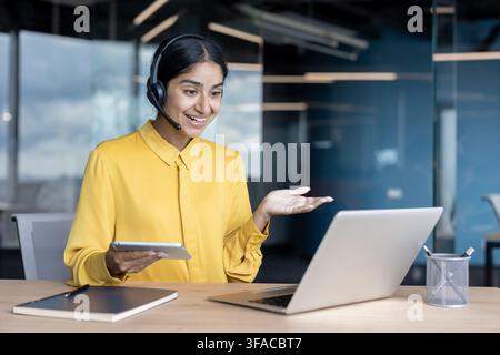 Jeune femme souriante avec casque, assise au bureau avec une tablette et discutant lors d'un appel vidéo via ordinateur portable. Banque D'Images