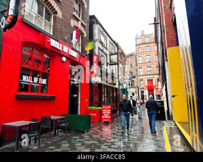 Liverpool, Royaume-Uni - 16 mai 2026 : Des piétons déambulent le long d'une ruelle pavée sous la pluie, devant des pubs, des enseignes au néon et des vitrines du quartier Cavern de Liverpool. Banque D'Images
