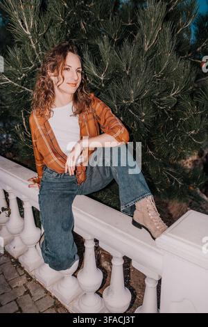 Femme nature en flanelle jeune femme joyeuse portant une chemise à carreaux et un jean en denim assise sur une rambarde en pierre près d'un pin vert Banque D'Images