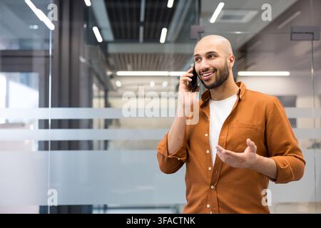 Homme professionnel ayant une conversation joyeuse au téléphone portable dans un cadre de travail moderne, communiquant activement et gesticulant avec une expression positive Banque D'Images