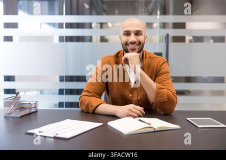 Homme chauve et confiant avec une barbe souriant, regardant la caméra et posant avec une main sous le menton tout en étant assis à un bureau dans un environnement de bureau moderne Banque D'Images