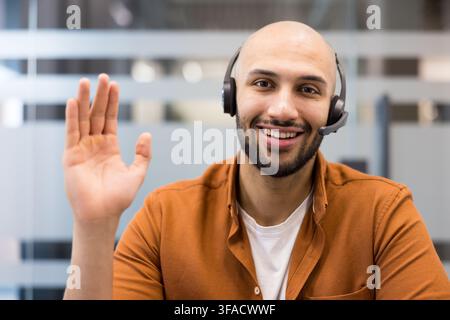 Homme chauve amical avec une barbe portant un casque et souriant à la caméra tout en agitant la main, fournissant un excellent service client dans un environnement de bureau professionnel Banque D'Images