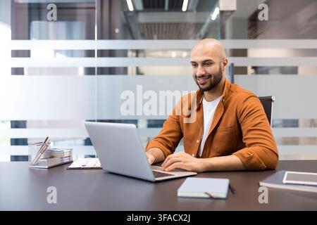 Jeune homme d'affaires travaillant joyeusement à son bureau contemporain, tapant sur un ordinateur portable et trouvant le succès, illustrant la productivité, la concentration et l'engagement dans un environnement d'entreprise Banque D'Images