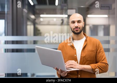 Jeune homme chauve debout avec assurance dans un cadre de bureau contemporain, tenant un ordinateur portable et regardant la caméra, symbolisant la technologie, le professionnalisme et l'entrepreneuriat Banque D'Images
