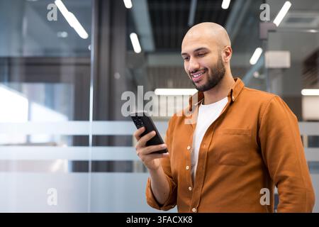 Jeune homme chauve avec une barbe souriant tout en tenant et interagissant avec un téléphone mobile dans un espace de travail contemporain, représentant la communication numérique et le réseautage Banque D'Images
