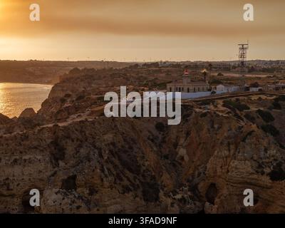 Vue aérienne du phare de Ponta da Piedade juché sur des falaises abruptes surplombant l'océan Atlantique lors d'un coucher de soleil chaud et doré à Faro Distri Banque D'Images
