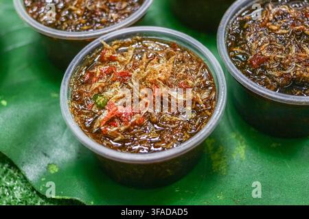 Une sauce poisson thaïlandaise sucrée avec des crevettes séchées et des tranches de piment repose dans des gobelets en plastique transparent sur un plateau à motifs verts, vue rapprochée. Banque D'Images