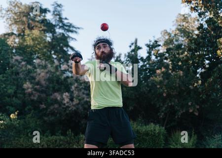 Homme jonglant habilement une balle rouge en plein air, démontrant un comportement concentré et actif dans un cadre naturel avec des arbres en arrière-plan, portant des gants noirs et une chemise verte, utilisation. Banque D'Images