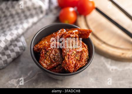 Tomates séchées au soleil empilées dans un bol sombre à côté de tomates cerises fraîches sur une planche en bois, évoquant les saveurs méditerranéennes, la cuisine rustique, les ingrédients conservés Banque D'Images