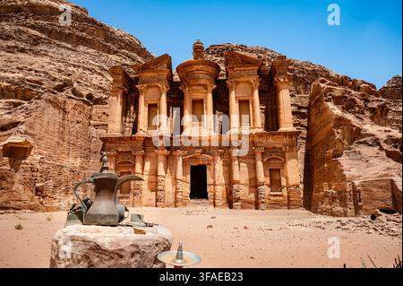 Caféière en laiton traditionnelle bedouine connue sous le nom de dallah posée sur une pierre au premier plan devant le monastère monumental Ad Deir taillé dans le grès Banque D'Images