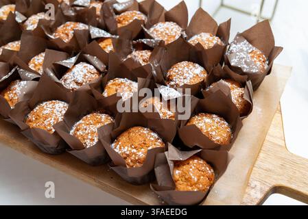 Un grand lot de muffins dorés, chacun saupoudré de sucre glace et enveloppé dans du papier brun foncé, disposés avec soin sur un plateau de service en bois Banque D'Images