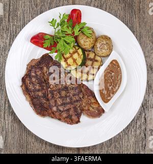 Steaks grillés, pommes de terre rôties et légumes sur une assiette blanche sur fond en bois Banque D'Images