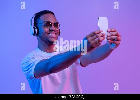 Beau Black Guy dans les casques sans fil et lunettes de soleil prenant Selfie Sur un smartphone Banque D'Images
