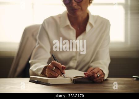 Femme d'affaires caucasienne d'âge moyen écrivant dans un carnet au bureau, planifiant un emploi du temps. Image adaptée à la communication d'entreprise, à la gestion du temps et aux supports de formation professionnelle Banque D'Images