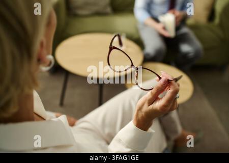 Thérapeute caucasienne d'âge moyen tenant des lunettes lors d'une consultation avec un patient masculin. L'image représente un soutien en santé mentale et est adaptée pour le marketing d'une clinique de psychologie Banque D'Images