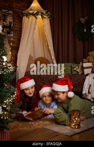 trois enfants portant des chapeaux du Père Noël, allongés sur un tapis et lisant ensemble un livre d'histoires à côté d'un sapin de Noël illuminé et de cadeaux emballés dans un Noël douillet Banque D'Images