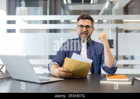 Homme d'affaires heureux applaudissant avec un coup de poing, tenant une enveloppe brune ouverte avec des documents, célébrant un succès professionnel, une promotion ou une offre d'emploi à un poste de travail moderne avec un ordinateur portable Banque D'Images