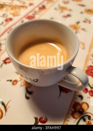 Café espresso frais dans une tasse en céramique blanche avec une crème dorée riche, servi sur une nappe à motif floral coloré, vue en angle Banque D'Images