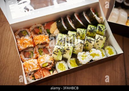Assortiment de sushi japonais servi dans une boîte à emporter, comprenant des rouleaux maki, des rouleaux de concombre, des sushi garnis de roe de poisson tobiko orange et des nigiri aux crevettes. Banque D'Images