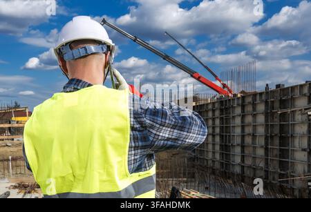 Un ouvrier du bâtiment observe l'avancement d'un projet de construction. Il porte un casque de sécurité et un gilet de sécurité. Une grue soulève des matériaux au Banque D'Images