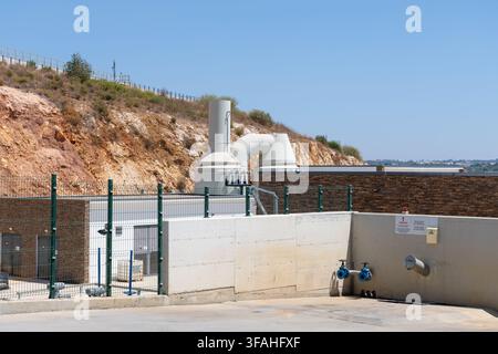 Une usine de traitement de l'eau moderne présente de gros tuyaux industriels, des murs en béton et des clôtures sur fond de colline rocheuse et de ciel bleu clair Banque D'Images