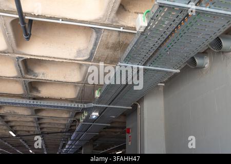 Un réseau complexe de bacs à câbles métalliques et de tuyaux court le long du plafond en béton, avec des tuyaux de ventilation sortant du mur Banque D'Images