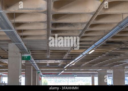 Une vue vers le haut d'une structure de plafond en béton moderne présentant des poutres apparentes et un éclairage industriel, avec des câbles et des tuyaux qui courent le long Banque D'Images