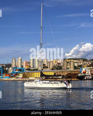 Yacht à voile blanc naviguant sur des eaux calmes du port de Rijeka avec un fond urbain et un ciel bleu, région de Kvarner, Croatie. Banque D'Images
