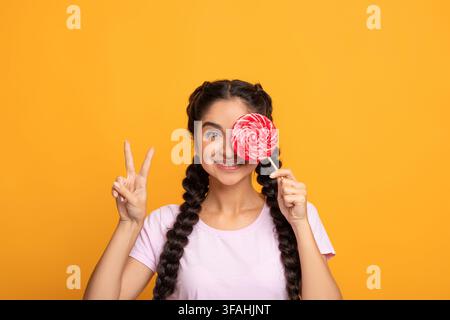 Femme indienne tenant un lollipop, couvrant l'œil, montrant le signe de la paix Banque D'Images