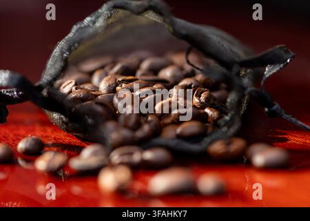 Des grains de café s'échappent d'un petit sachet en filet noir sur une surface rouge. Banque D'Images