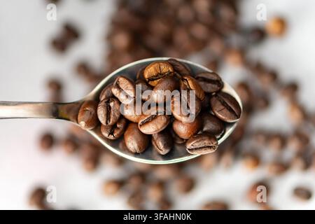Une cuillère en métal tient des grains de café torréfiés sur un fond blanc. Banque D'Images