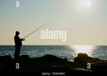 Silhouette d'un pêcheur solitaire debout sur une digue rocheuse tenant une canne à pêche contre une mer baignée de soleil. L'homme lance sa ligne dans Banque D'Images