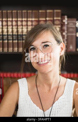 Portrait rapproché d'une jeune femme souriante avec des lunettes regardant au loin dans une bibliothèque à domicile, exprimant la confiance, l'optimisme, le bien-être et une vie positive Banque D'Images