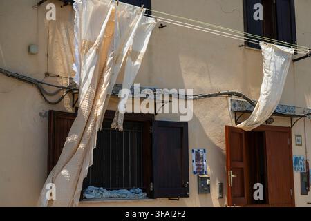 Prise de vue détaillée de textiles traditionnels en dentelle blanche flottant doucement sur une corde à linge devant un mur en plâtre beige chaud et une fenêtre en bois ouverte à Mondello. Banque D'Images