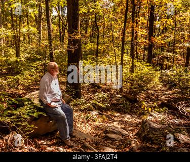 Un homme mûr est assis sur un rocher dans une forêt automnale baignée de soleil au Gambrill State Park à Frederick, dans le Maryland, prenant une pause tranquille entouré d'arbres et d'automne Banque D'Images