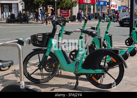 Une rangée de vélos électriques verts Bolt garés sur un trottoir à Liverpool, Royaume-Uni, avec des rues de la ville et des piétons en arrière-plan. Banque D'Images