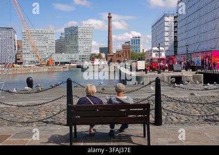 Vue arrière d'un couple de personnes âgées assis sur un banc au Royal Albert Dock à Liverpool, Royaume-Uni, regardant au loin sur l'eau vers le pub The Pump House Banque D'Images