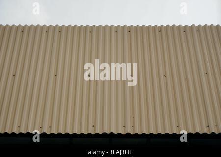 Vue rapprochée d'un panneau de toit en métal ondulé beige devant un ciel pâle et lumineux. Une texture architecturale minimaliste présentant des stries verticales. Banque D'Images