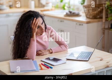 Femme indépendante ennuyeuse assise à la table avec ordinateur portable dans la cuisine Banque D'Images