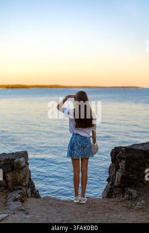 Femme regardant à travers l'eau calme au coucher du soleil. Femme debout sur une rive rocheuse, regardant à travers l'eau bleue calme vers l'horizon au coucher du soleil. Banque D'Images