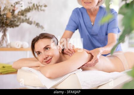 Femme mature recadrée frotte activement l'épaule de la jeune femme pendant la procédure générale de massage. Banque D'Images