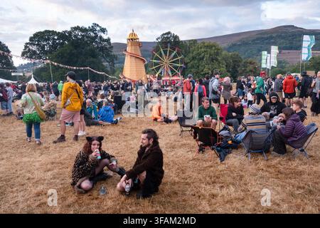 Brecon, Pays de Galles, Royaume-Uni, 20 août 2026. Au premier plan, un jeune couple vêtu de manière extravagante est assis sur l'herbe, tandis que des foules de personnes se détendent dans un grand champ avec une attraction foraine derrière eux. Photo : Rob Watkins/Alamy Live News. INFO : Après des mois de sécheresse, de fortes averses sont tombées le premier jour du Green Man Festival 2026 et la foule a apprécié le changement rafraîchissant de la météo et s'est également mise à l'abri dans les tentes de l'arène. Banque D'Images