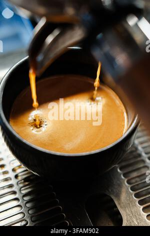 Extraction d'espresso en double flux versant dans une tasse en céramique sombre remplie d'une épaisse crème dorée. Prise de vue professionnelle d'une machine à café. Banque D'Images
