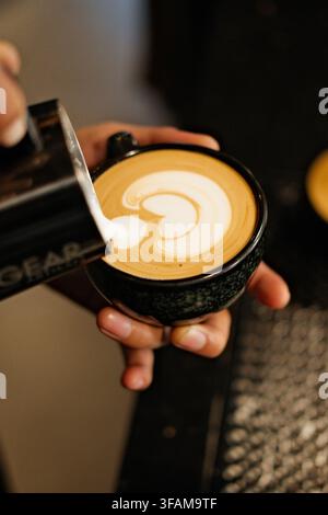 Barista versant du lait moussé dans une tasse en céramique noire pour créer un art latte tulipe. Gros plan de la préparation d'un café spécial dans un café. Banque D'Images