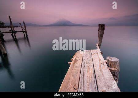 Quai en bois patiné s'étendant sur une eau sombre et calme vers des montagnes brumeuses sous un ciel d'aube doux et violet, créant une scène tranquille et paisible au bord du lac Banque D'Images