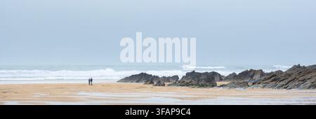 Une image panoramique d'un couple debout près de rochers exposés par une marée basse sur Fistral Beach par un temps brumeux et froid à Newquay, Cornwall, au Royaume-Uni. Banque D'Images