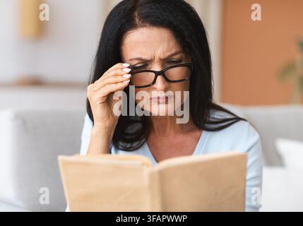Femme mûre confuse dans les verres essayant de lire le livre Banque D'Images