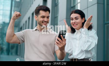 Deux collègues d'affaires caucasiens excités, heureux et choqués gagnant avec un téléphone portable et se donnant un high five en ville. Homme positif et émerveillé avec une femme d'affaires Banque D'Images