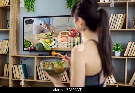 Femme en tenue de sport mangeant une salade tout en regardant un tutoriel de cuisine à la télévision à la maison Banque D'Images